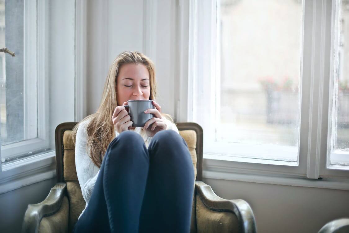 A woman with eyes closed sitting in a vintage armchair by a window, holding a grey mug to her face, highlighting traditional medicine approaches to managing long COVID.