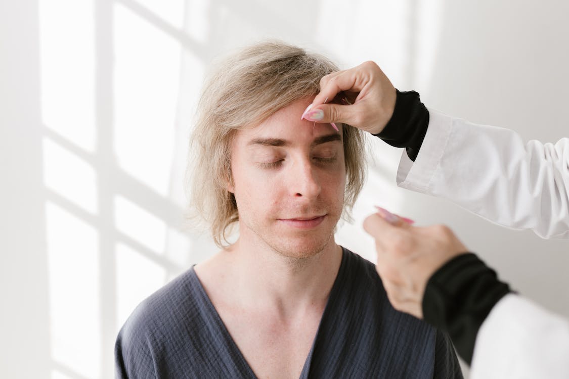 Practitioner in a white lab coat performing facial acupuncture on a relaxed blonde man, illustrating musculoskeletal pain relief with acupuncture and TCM.