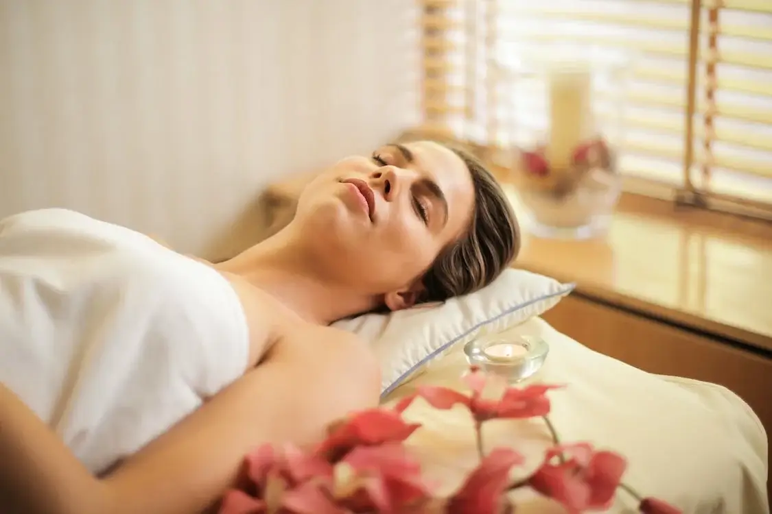 A woman lying on a massage table with her eyes closed in a serene spa setting decorated with pink flowers and a burning candle, promoting better sleep and treating insomnia with acupuncture and traditional Chinese medicine.