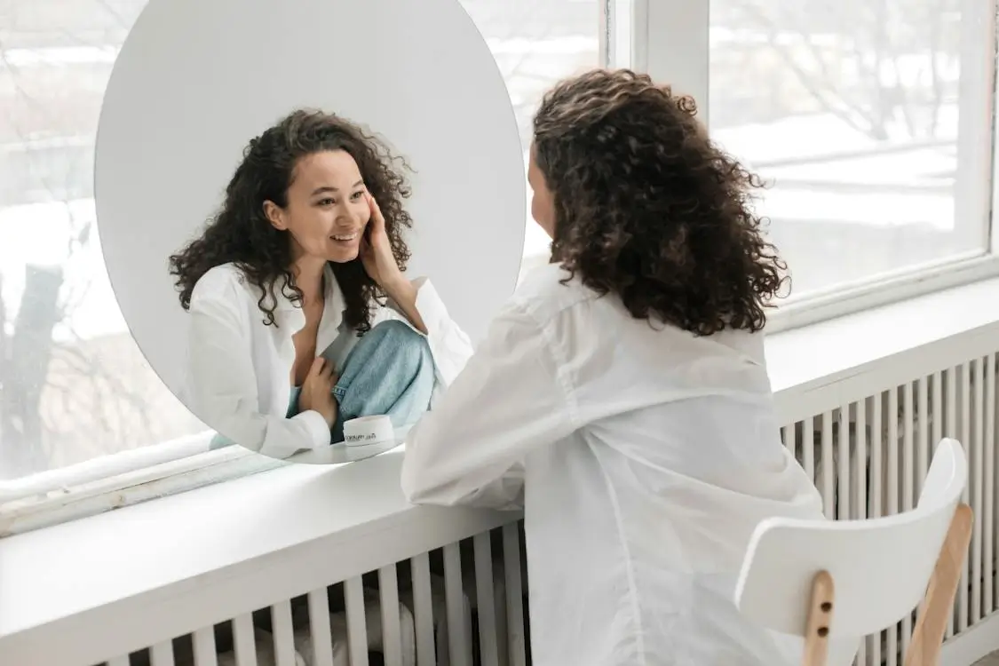A woman with curly hair wearing a white button-down shirt smiling while looking at her reflection in a round mirror and gently touching her face, promoting self-care and boosting self-love with Traditional Chinese Medicine.