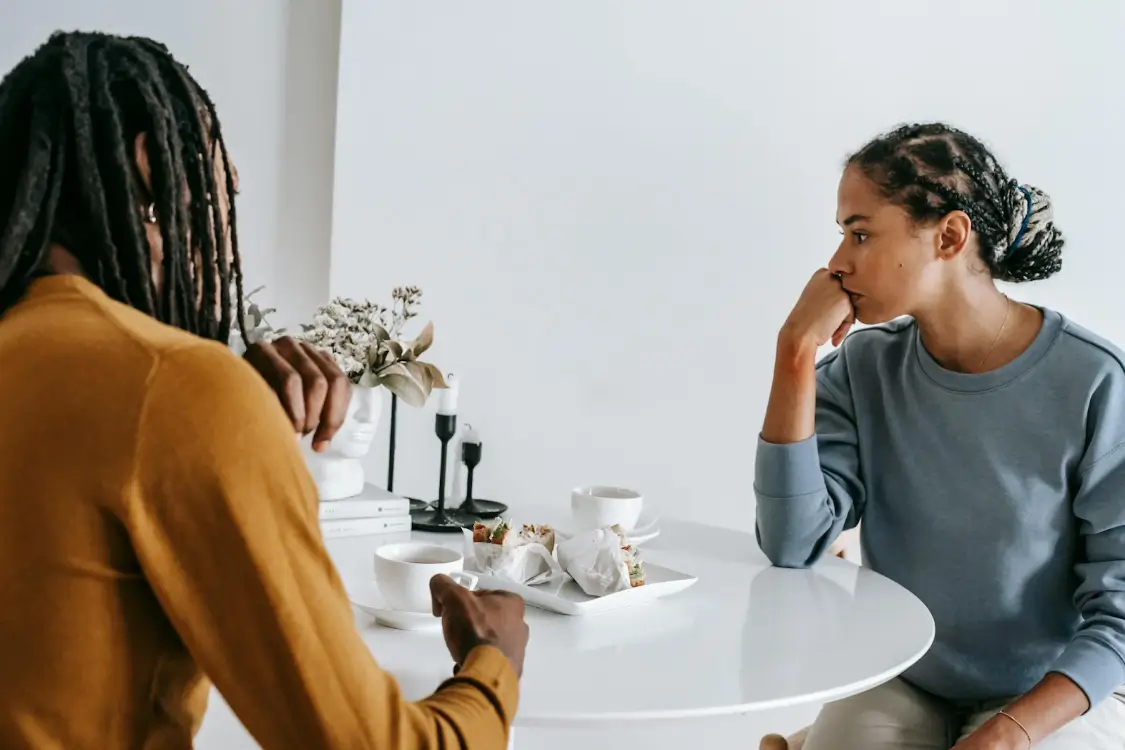 An image of two people with braided hair sitting at a white table with coffee and sandwiches, one looking away thoughtfully with their chin resting on their hand, illustrating natural remedies for anger and TCM tips on managing it at Miami Acupuncture and Classical Herbs.